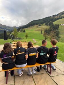 Jugendliche in T-Shirts mit Aufdruck sitzen auf Bank vor Bergkulisse in den Alpen.